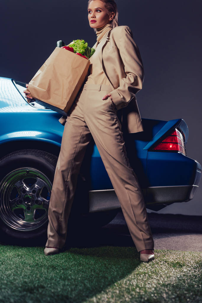 atrakcyjna i stylowa kobieta w garniturze stojąca w pobliżu samochodu retro i trzymająca papierową torbę z jedzeniem  - Zdjęcie, obraz