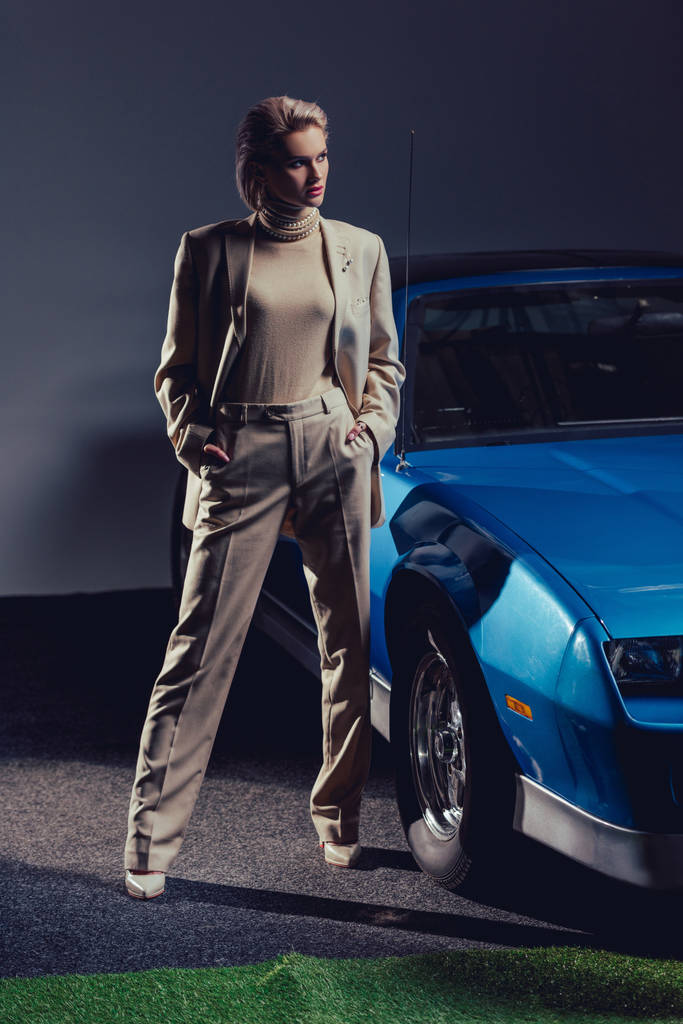 atrakcyjna i stylowa kobieta w garniturze z rękami w kieszeniach stojąca w pobliżu samochodu retro  - Zdjęcie, obraz