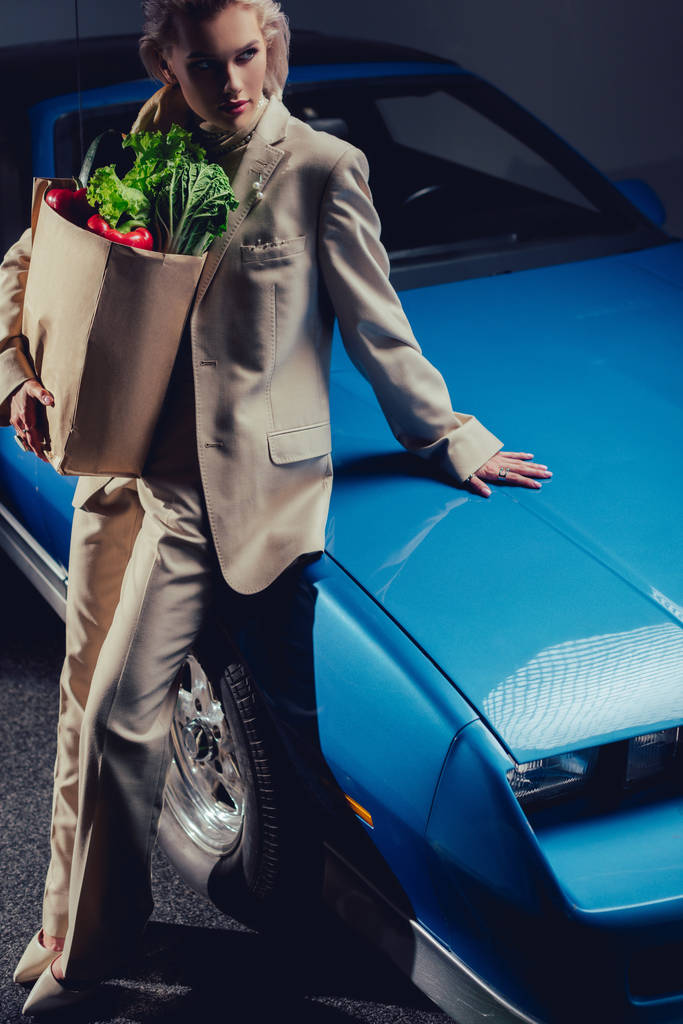レトロな車の近くに立つスーツ姿の魅力的でスタイリッシュな女性の高い角度ビューと食品と紙袋を保持 - 写真・画像