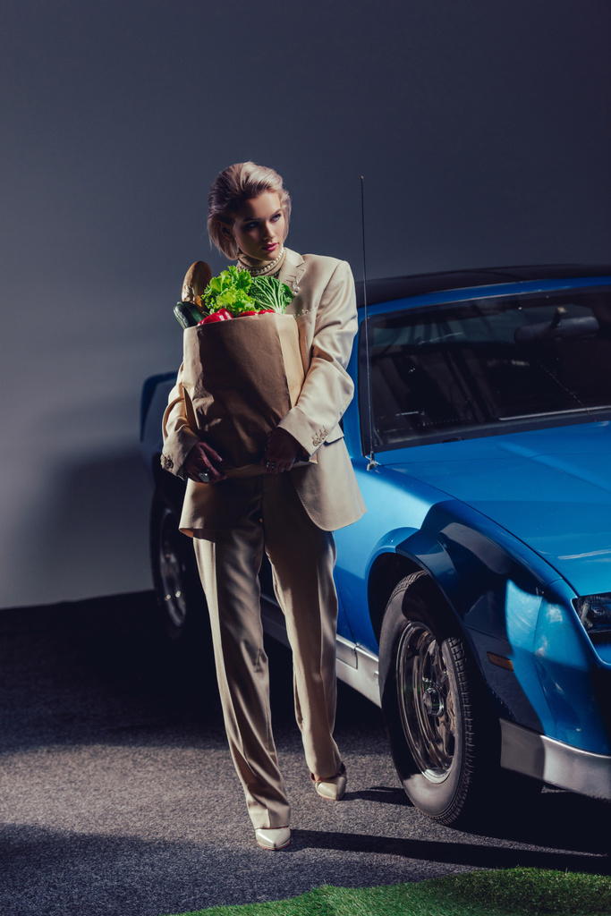 atraktivní a stylová žena v obleku stojí v blízkosti retro auta a drží papírový sáček s jídlem  - Fotografie, Obrázek