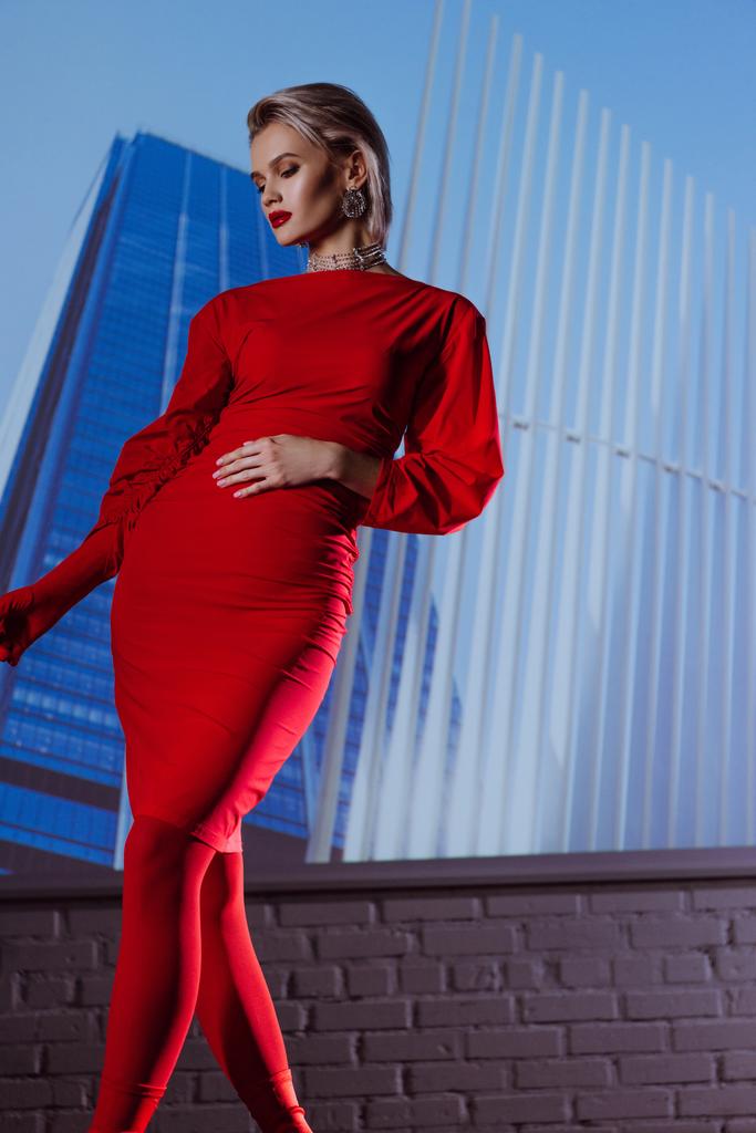 niski kąt widzenia atrakcyjnej i stylowej kobiety w czerwonej sukience na tle miasta  - Zdjęcie, obraz