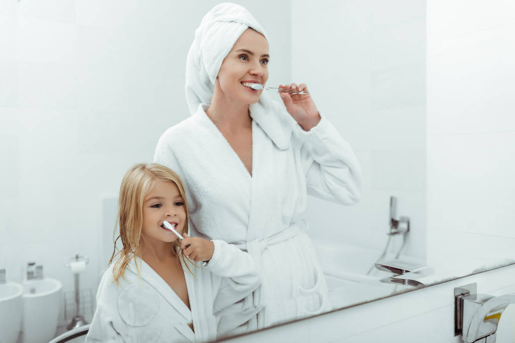 šťastná matka a dcera v župany čištění zubů v blízkosti zrcadla  - Fotografie, Obrázek