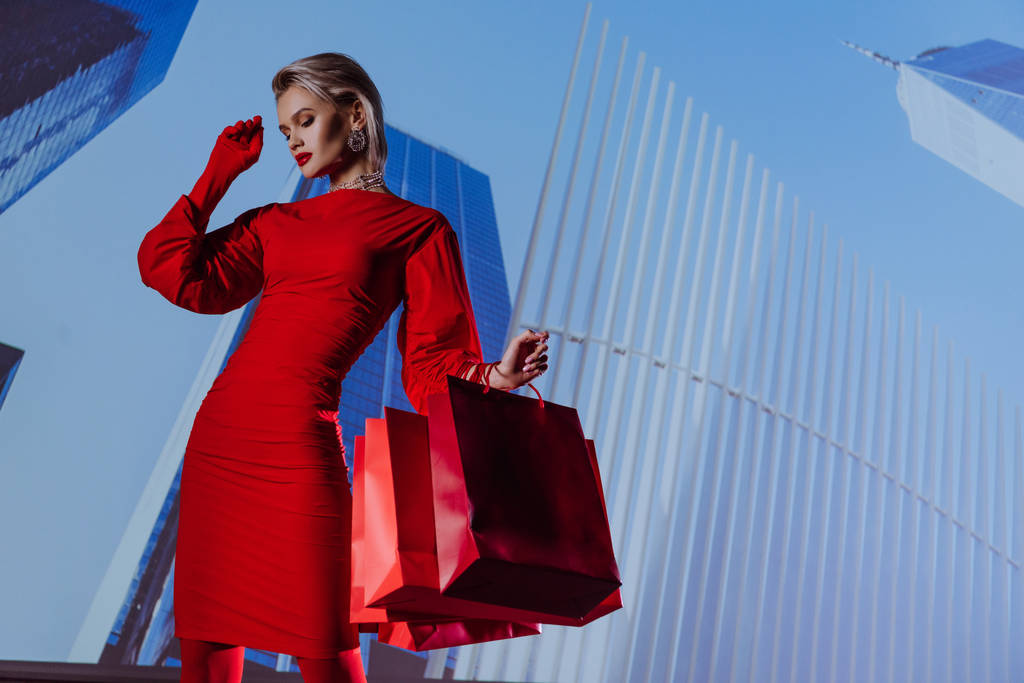 low angle view of attractive and stylish woman in red dress holding shopping bags on city background  - Photo, Image