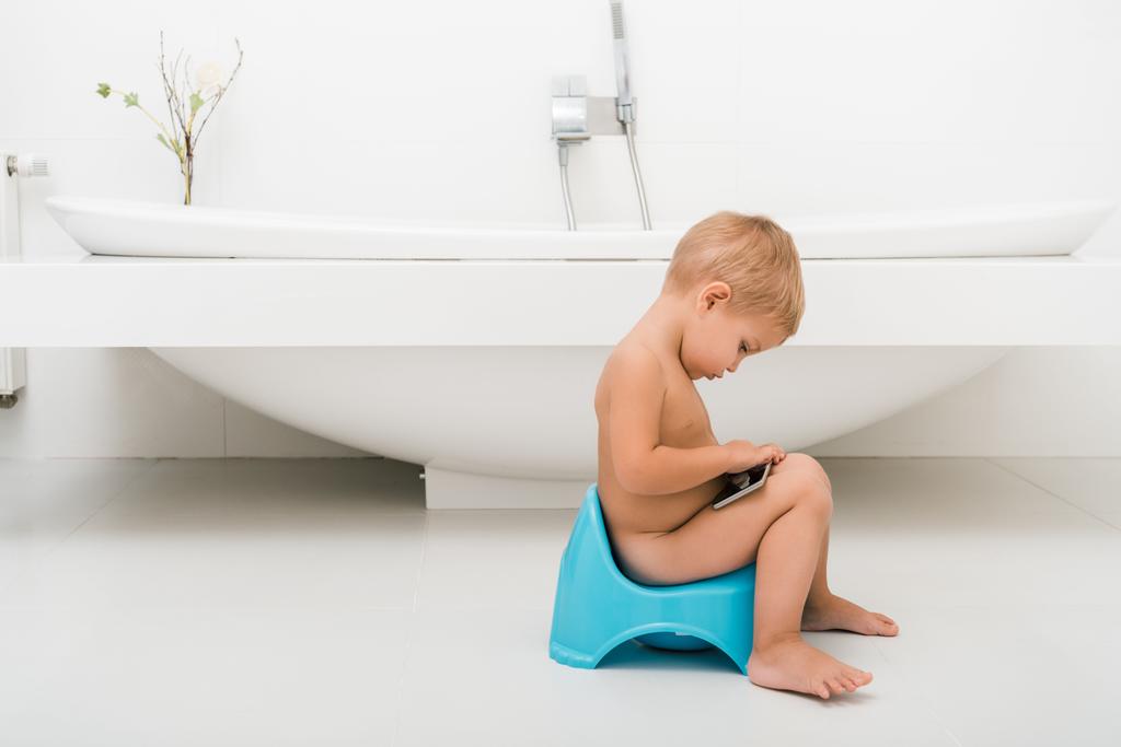 vue latérale du garçon tout-petit assis sur le pot bleu et tenant smartphone près de la baignoire
  - Photo, image