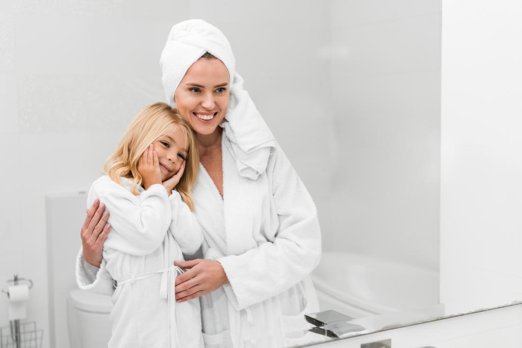 Χαρούμενη μητέρα και χαριτωμένη κόρη σε μπουρνούζια κοιτάζοντας καθρέφτη στο μπάνιο  - Φωτογραφία, εικόνα