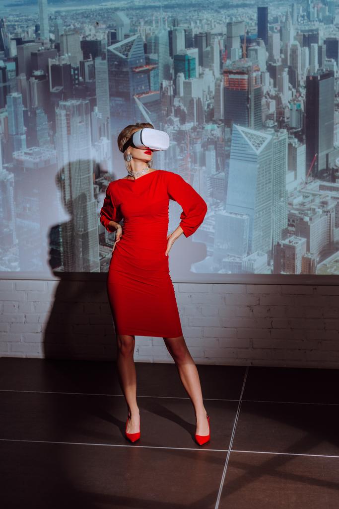 mujer elegante con las manos en las caderas en vestido rojo y auriculares de realidad virtual en el fondo de la ciudad
  - Foto, imagen