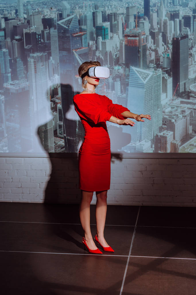 tyylikäs nainen ojennetut kädet punainen mekko ja virtuaalitodellisuus kuulokkeet kaupungin taustalla
  - Valokuva, kuva