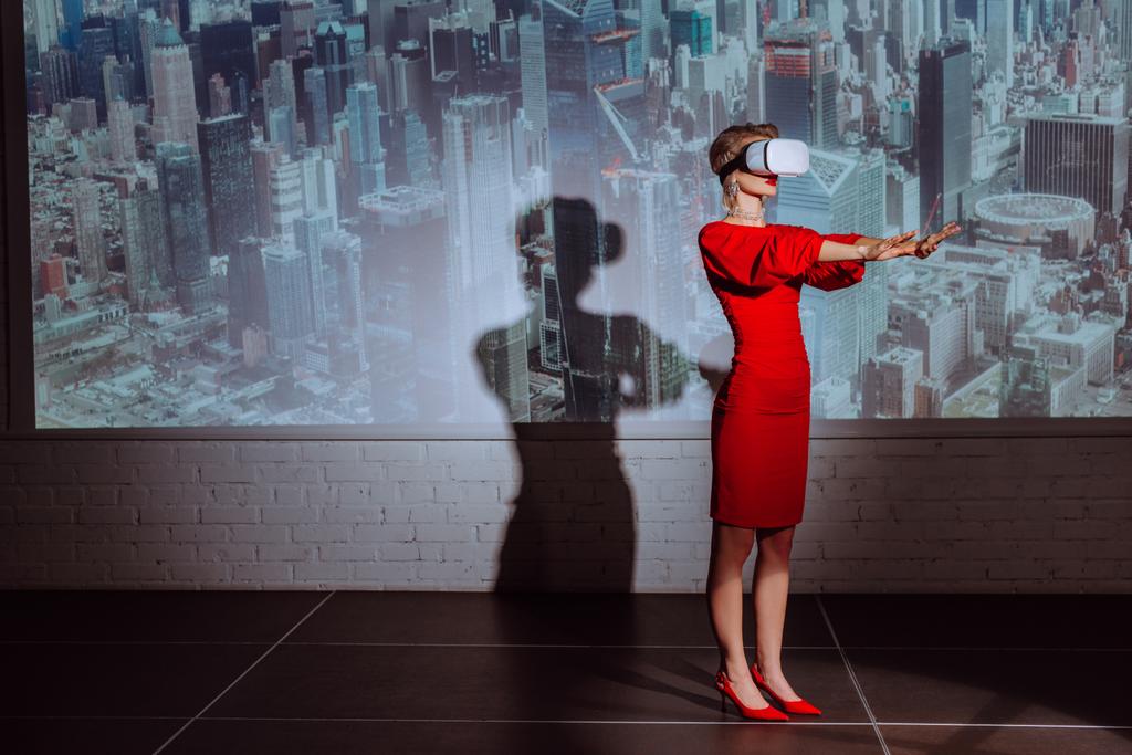 κομψή γυναίκα με απλωμένα χέρια σε κόκκινο φόρεμα και ακουστικά εικονικής πραγματικότητας στο φόντο της πόλης  - Φωτογραφία, εικόνα