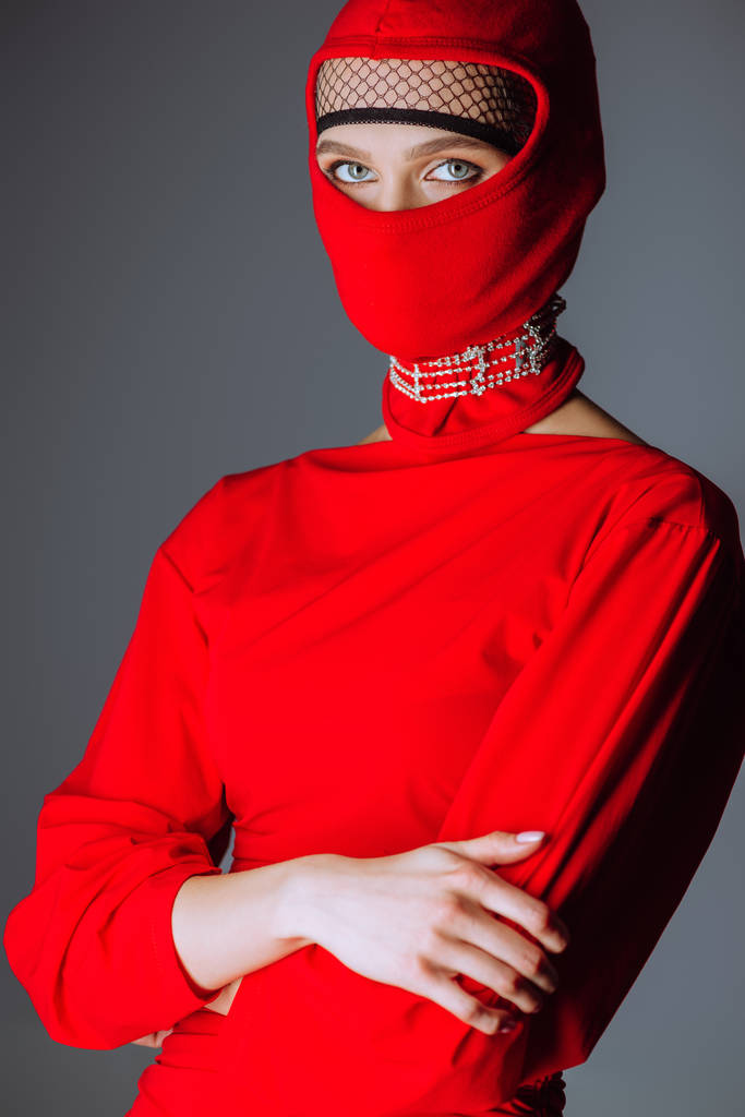 κομψή γυναίκα με κόκκινο φόρεμα και balaclava με σταυρωμένα χέρια απομονώνονται σε γκρι - Φωτογραφία, εικόνα