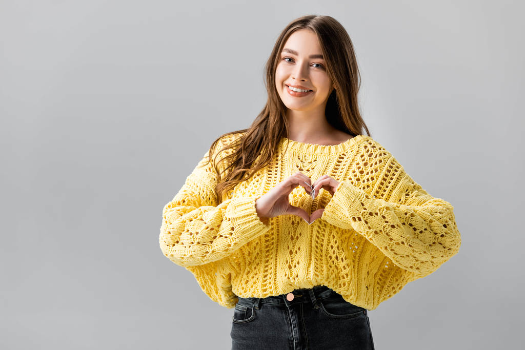 χαμογελαστό κορίτσι σε κίτρινο πουλόβερ δείχνει την καρδιά σημάδι απομονώνονται σε γκρι - Φωτογραφία, εικόνα