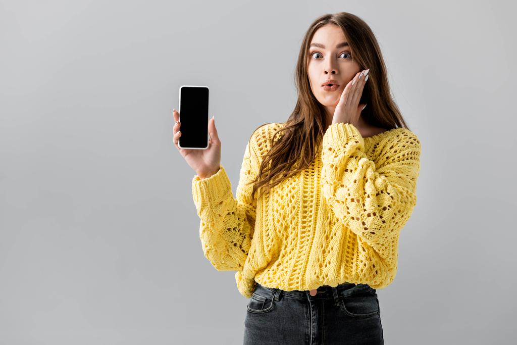 σοκαρισμένη κοπέλα συγκινητικό πρόσωπο, ενώ δείχνει smartphone με λευκή οθόνη απομονωμένη σε γκρι - Φωτογραφία, εικόνα