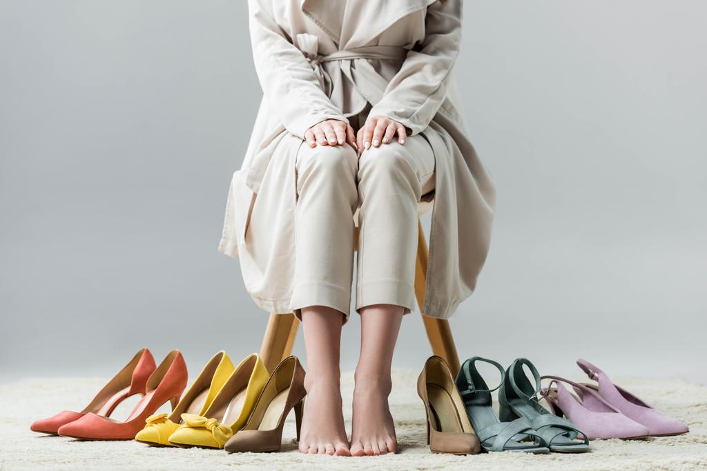 vista cortada de menina descalça sentada na cadeira perto da coleção de sapatos no fundo cinza
 - Foto, Imagem