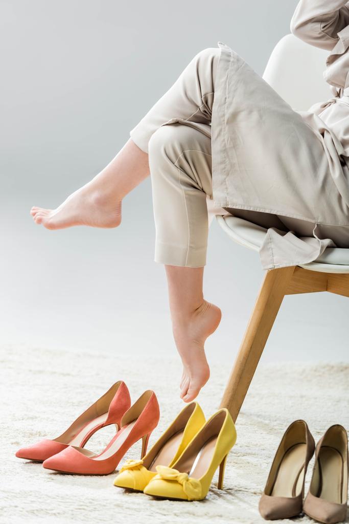cropped view of barefoot girl sitting on chair near collection of shoes on grey background - Photo, Image