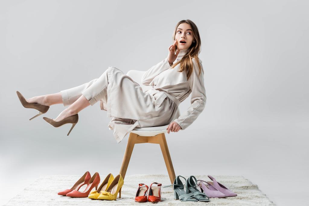 surprised girl looking away while sitting on chair with raised legs near collection of shoes on grey background - Photo, Image