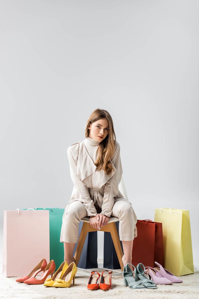 módní, sebevědomá dívka dívá na kameru, zatímco sedí na židli v blízkosti nákupních tašek a sbírka obuvi na šedém pozadí - Fotografie, Obrázek