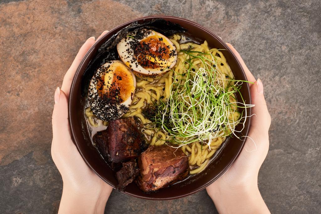 cropped view of woman holding bowl with spicy meat ramen with chopsticks on stone surface - Photo, Image
