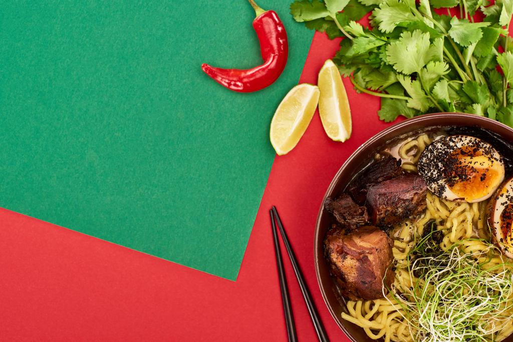 κορυφαία άποψη του πικάντικου ράμεν κρέατος κοντά σε φρέσκα υλικά και chopsticks σε πράσινη και κόκκινη επιφάνεια - Φωτογραφία, εικόνα