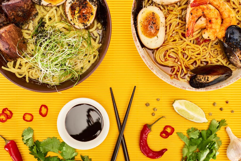 top view of seafood and meat ramen near fresh ingredients, soy sauce and chopsticks on yellow surface - Photo, Image