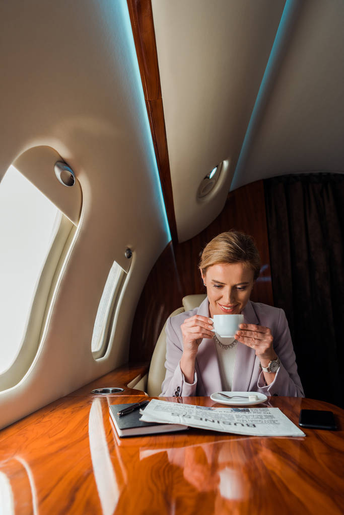 glückliche Geschäftsfrau hält Tasse in der Hand, während sie im Privatflugzeug Wirtschaftszeitung liest  - Foto, Bild