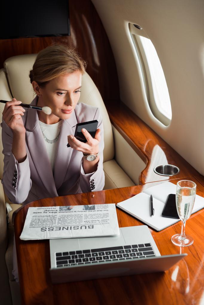 Frau trägt dekorative Kosmetik in der Nähe von Gadgets und Champagnergläsern im Privatflugzeug auf  - Foto, Bild