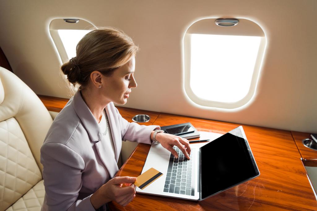 ελκυστική επιχειρηματίας που χρησιμοποιεί φορητό υπολογιστή με κενή οθόνη και κρατώντας πιστωτική κάρτα σε ιδιωτικό αεροπλάνο  - Φωτογραφία, εικόνα