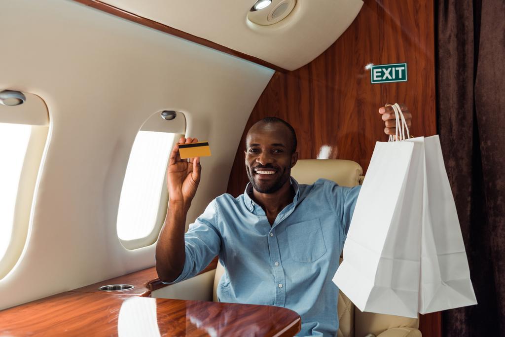 glücklicher afrikanisch-amerikanischer Mann mit Kreditkarte und Einkaufstaschen im Privatflugzeug  - Foto, Bild