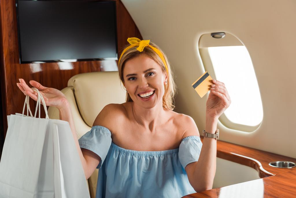 cheerful girl holding credit card and shopping bags in private plane  - Photo, Image