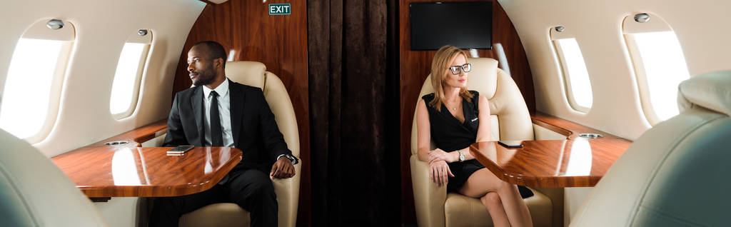 panoramiczne ujęcie przystojnego afrykańskiego biznesmena i atrakcyjnej bizneswoman siedzi w prywatnym samolocie  - Zdjęcie, obraz