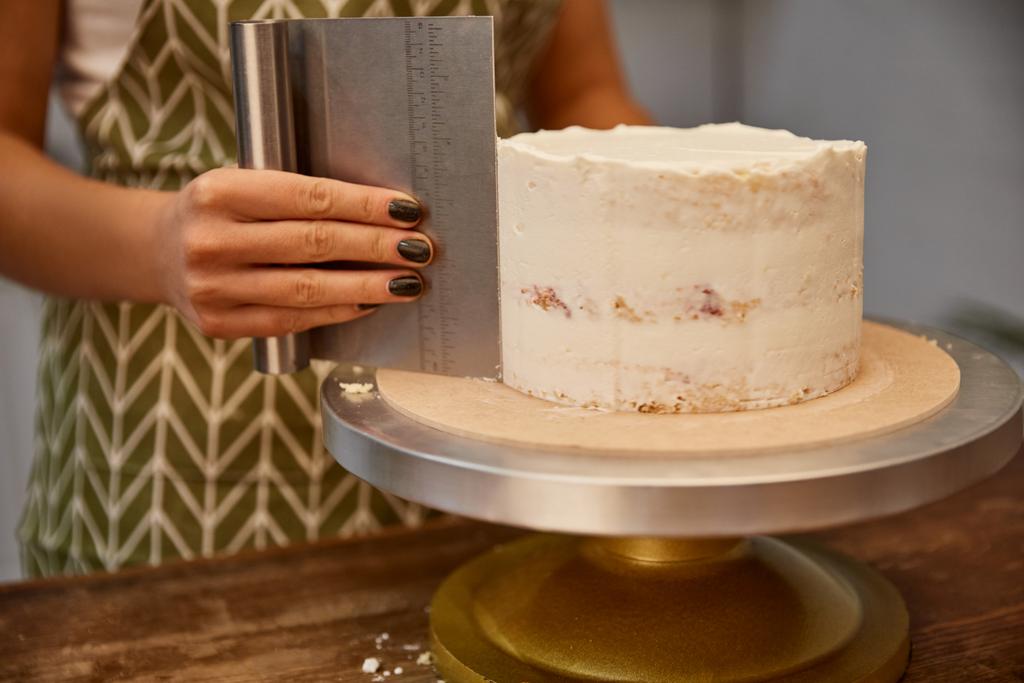 Vista recortada del confitero usando raspador en crema sabrosa en la torta de esponja
  - Foto, Imagen
