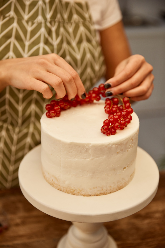 Vista recortada del confitero poniendo grosella roja jugosa en la torta con crema
 - Foto, Imagen