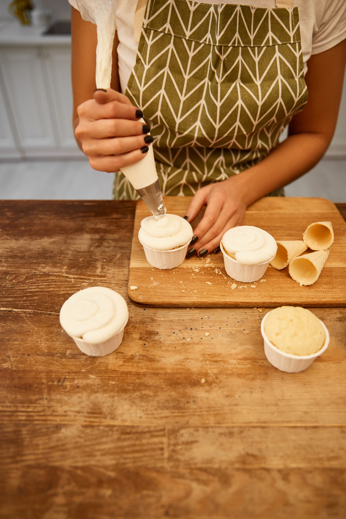Обрезанный вид кондитера, добавляющего сладкий крем на кексы рядом с вафельными рожками на разделочной доске
 - Фото, изображение