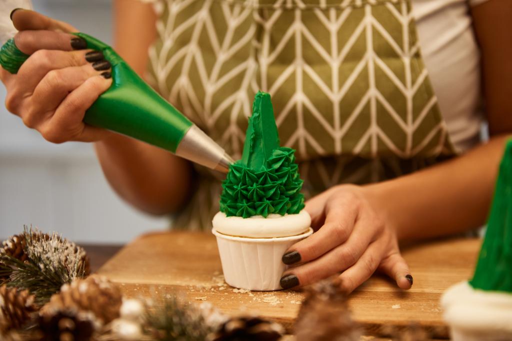 Cropped view of confectioner decorating Christmas tree cupcake with green cream on table with spruce cones  - 写真・画像
