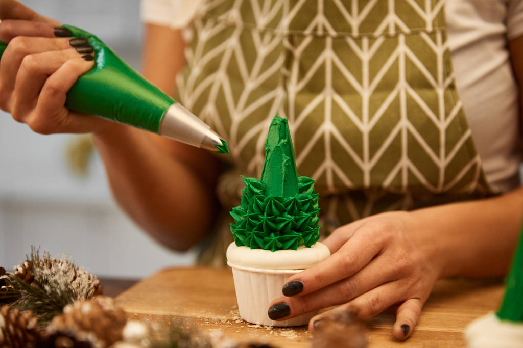 Vue recadrée du confiseur ajoutant de la crème verte sur le cupcake du sapin de Noël à côté des cônes d'épinette sur la table
 - Photo, image