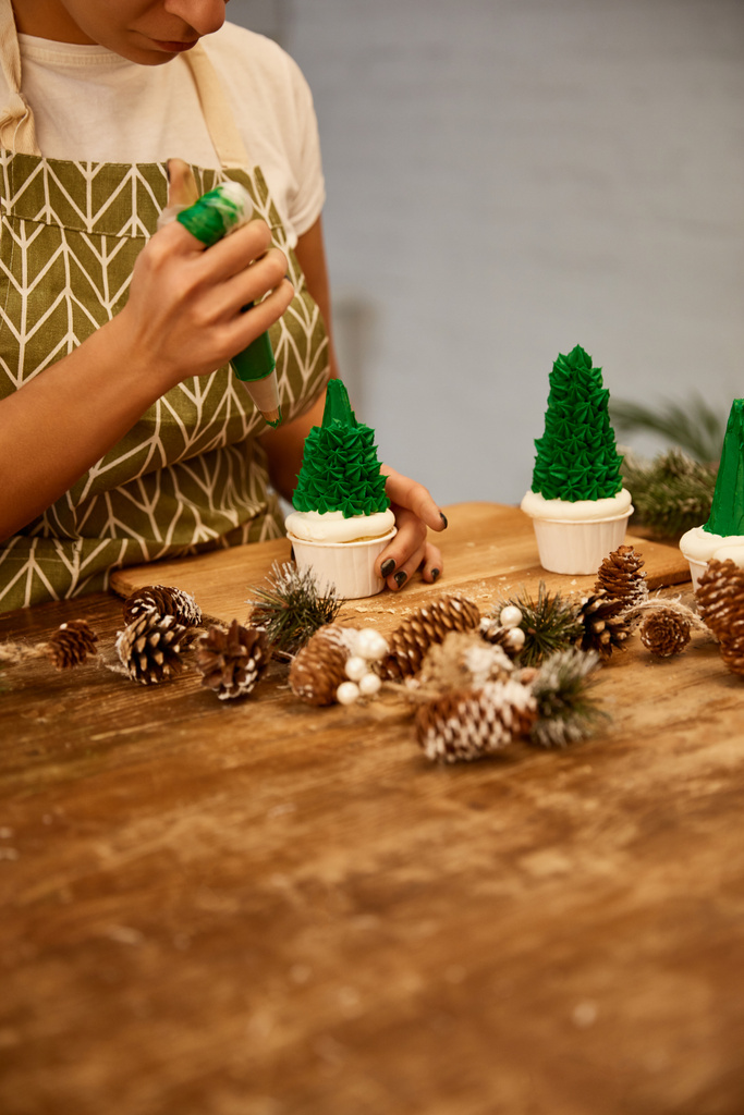 Oříznutý pohled na cukráře zdobení vánoční stromeček cupcakes se zelenou sladkou smetanou vedle smrkových kuželů na stole - Fotografie, Obrázek