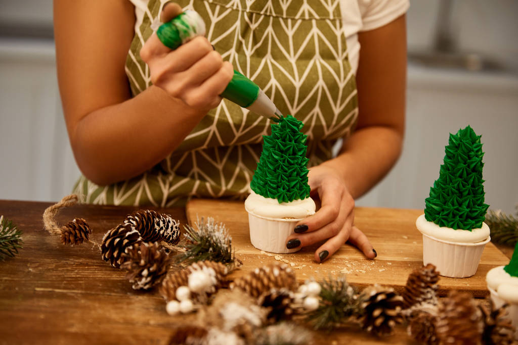 Gekropte weergave van banketbakker toevoegen van groene zoete room op kerstboom cupcakes met sparren kegels op tafel - Foto, afbeelding
