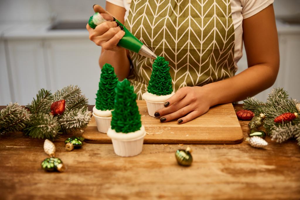 zugeschnittene Ansicht eines Konditors, der Weihnachtsbaum-Cupcakes neben Fichtenzweigen und Weihnachtsdekor auf dem Tisch herstellt - Foto, Bild