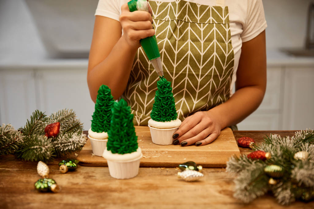 Vista recortada del pastelero con bolsa de pastelería añadiendo crema en cupcakes de árbol de Navidad con ramas de pino en la mesa
 - Foto, imagen