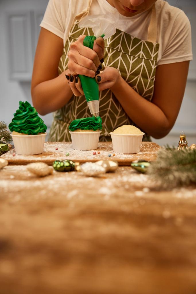 zugeschnittene Ansicht eines Konditors, der mit grüner Sahne und Cupcakes neben Kugeln und Tannenzweigen auf dem Tisch arbeitet - Foto, Bild