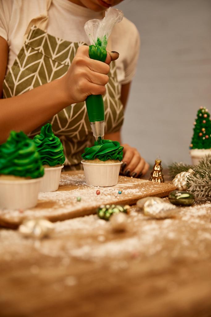 Gekropte weergave van banketbakker werkend met room en cupcakes naast kerstballen en dennentak op tafel - Foto, afbeelding