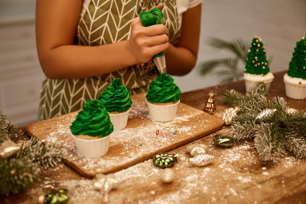 Vue recadrée du confiseur décorant les cupcakes du sapin de Noël à côté des branches d'épinette et des décorations de Noël sur la table
 - Photo, image