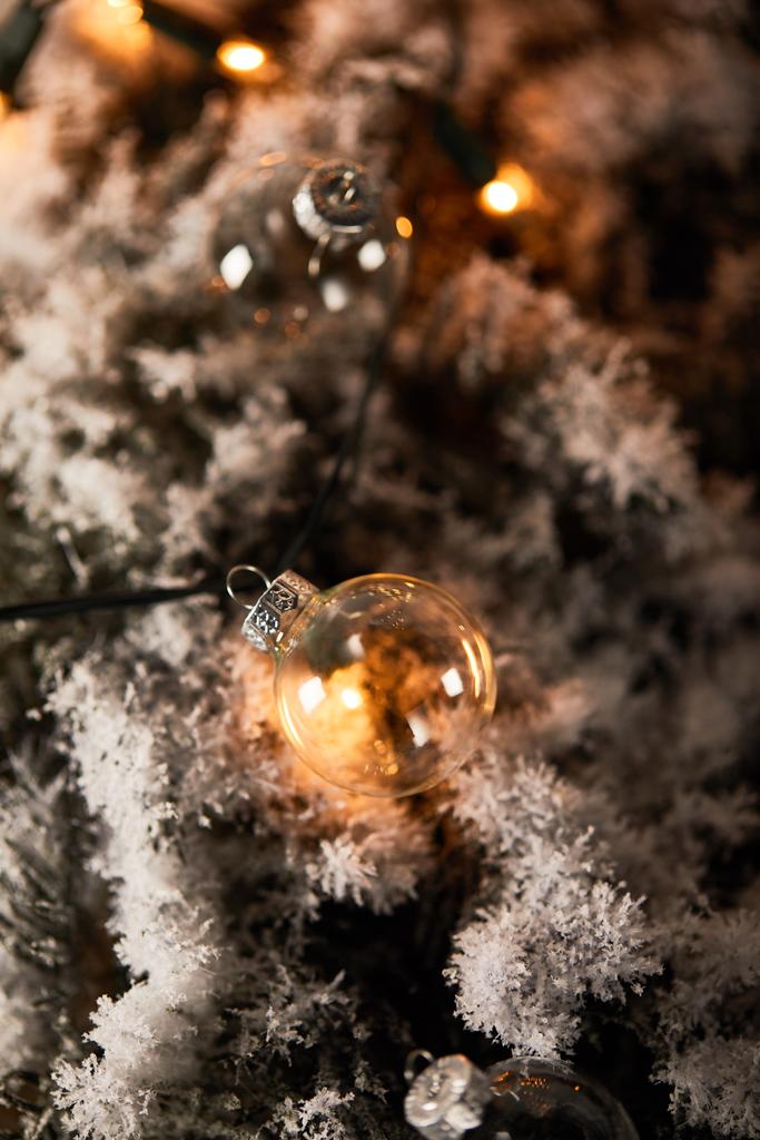 Selektiver Fokus von transparenten Weihnachtskugeln und gelben Lichtern auf Fichtenzweigen im Schnee - Foto, Bild