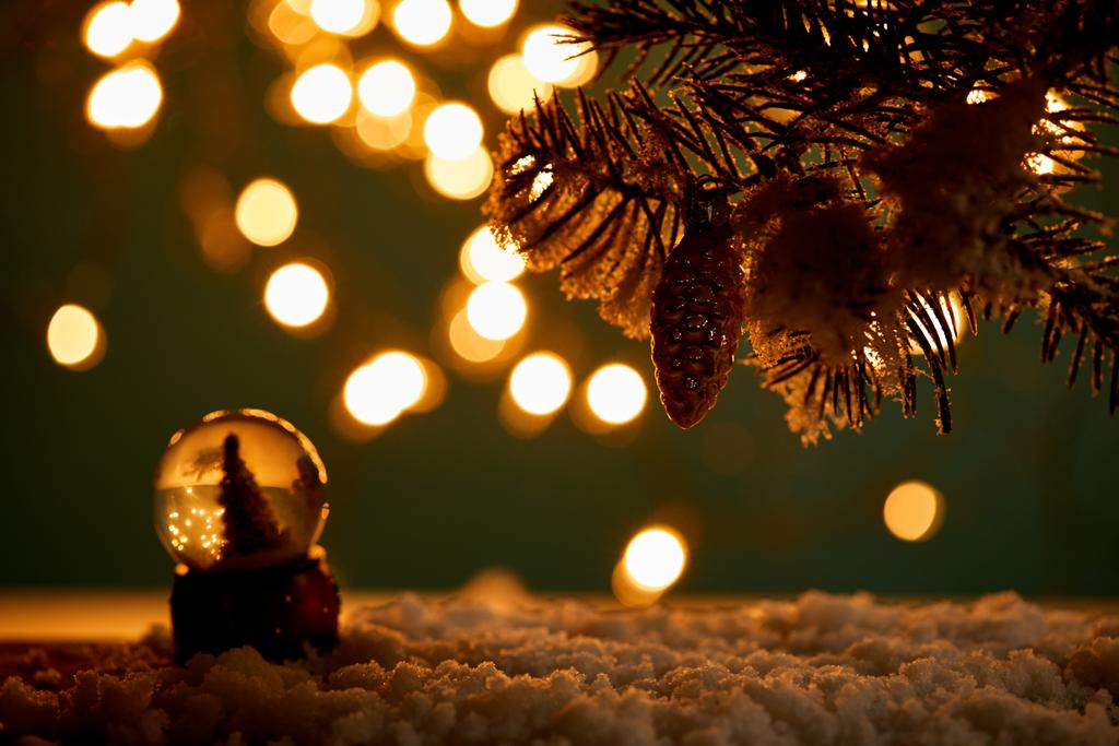 mała kula śnieżna z choinką stojącą w śniegu z gałązkami świerku, kulą świąteczną i rozmazanymi światłami w nocy  - Zdjęcie, obraz