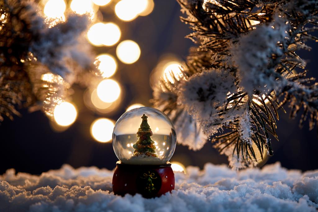 arbre de Noël décoratif en boule de neige debout dans la neige avec des branches d'épinette et des lumières floues la nuit
 - Photo, image