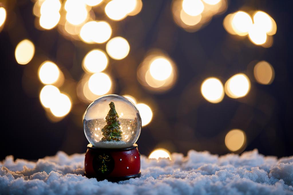 χριστουγεννιάτικο δέντρο σε χιονόμπαλα στέκεται στο χιόνι με θολή φώτα τη νύχτα - Φωτογραφία, εικόνα