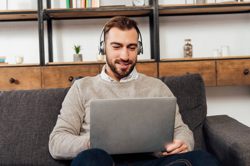 Sorrindo homem em fones de ouvido usando laptop no sofá na sala de estar
 - Foto, Imagem