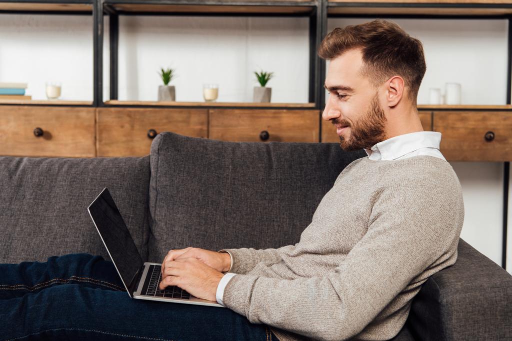 Homme souriant tapant sur un clavier d'ordinateur portable sur un canapé dans le salon
 - Photo, image