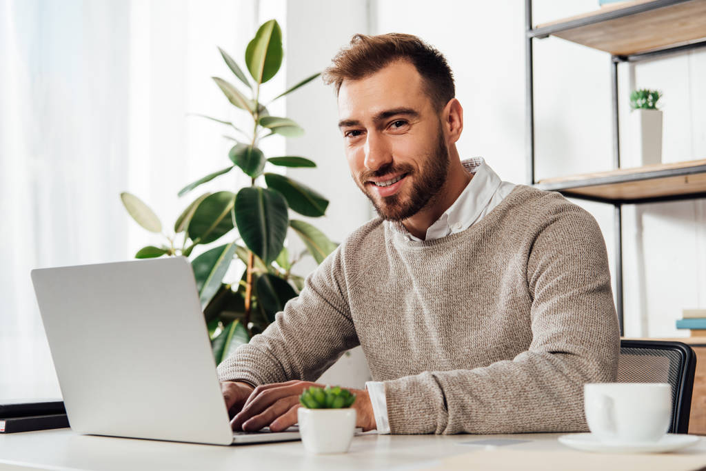 Smiling man looking at camera while using laptop at home - Photo, Image