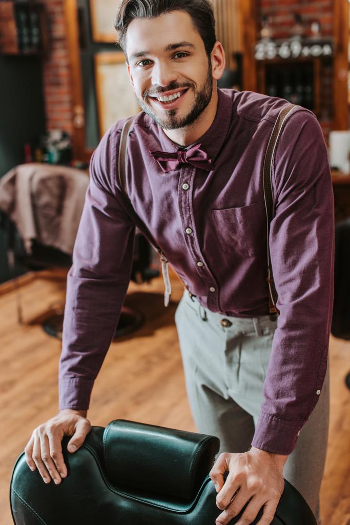 cheerful bearded barber touching leather armchair in barbershop  - Photo, Image