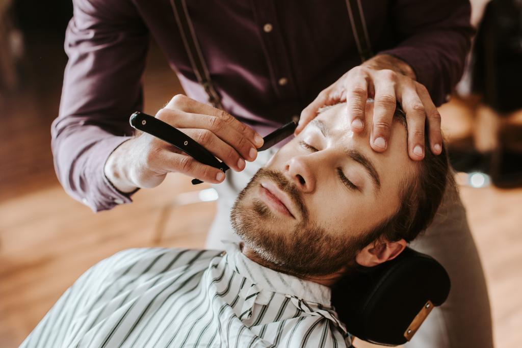 Selektiver Fokus des Barbiers rasiert bärtigen Mann mit geschlossenen Augen  - Foto, Bild