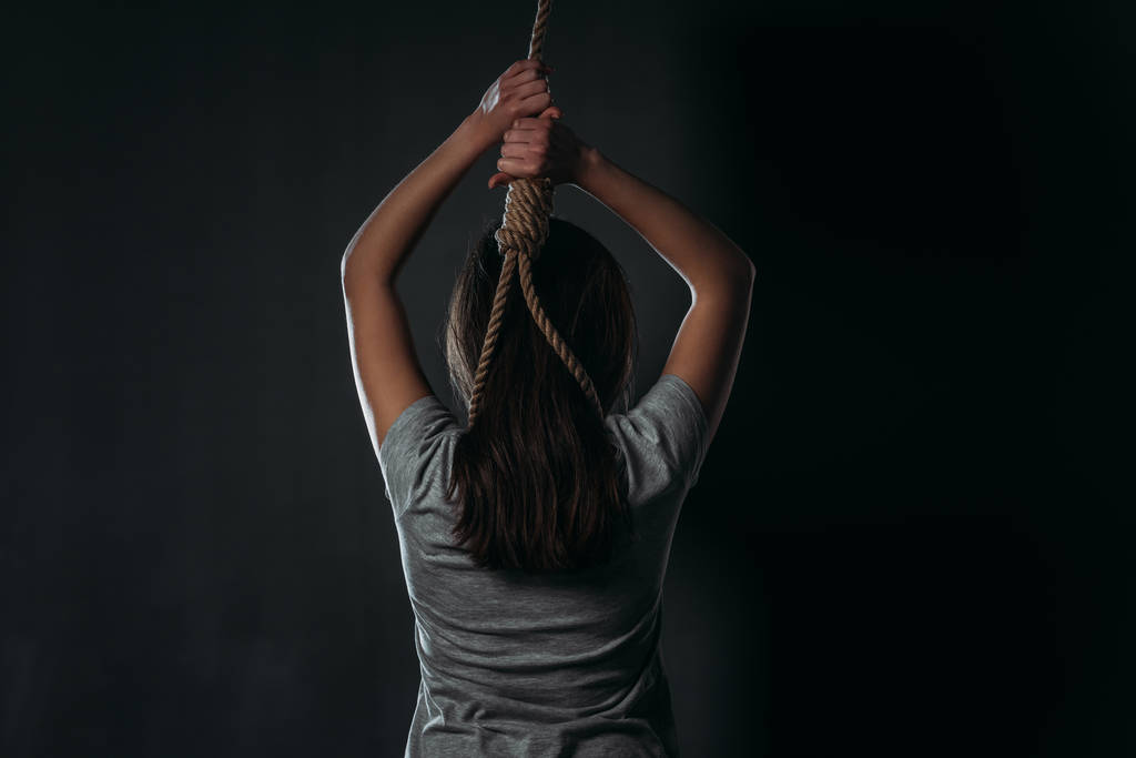 Rückansicht einer Frau, die Selbstmord begeht, während sie eine Schlinge um den Hals auf schwarzem Hintergrund legt - Foto, Bild